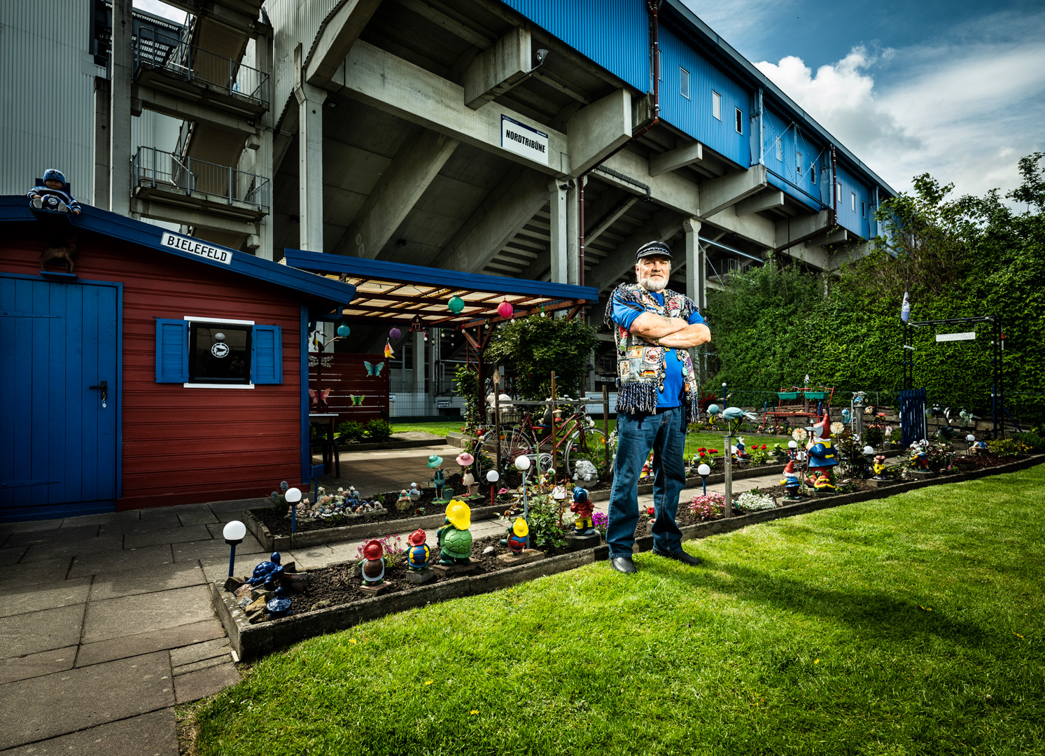 Ein Mann mit Jeansweste steht in seinem Schrebergarten. Ein Stadion im Hintergrund.