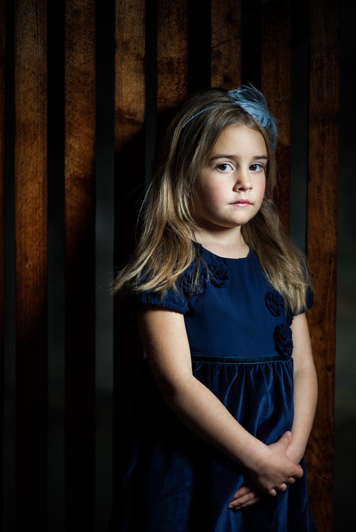 Ein Mädchen in einem blauen Kleid steht vor einer Holzwand.